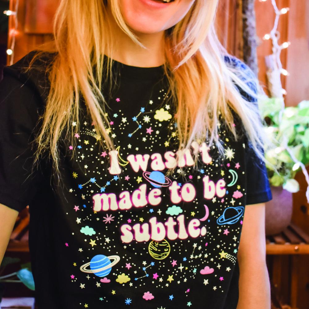 I Wasn’t Made to Be Subtle black graphic t-shirt with colorful planets, stars, and space-themed design, modeled by a smiling person with blonde hair – vibrant retro cosmic tee – The Funnel Cake Tree.