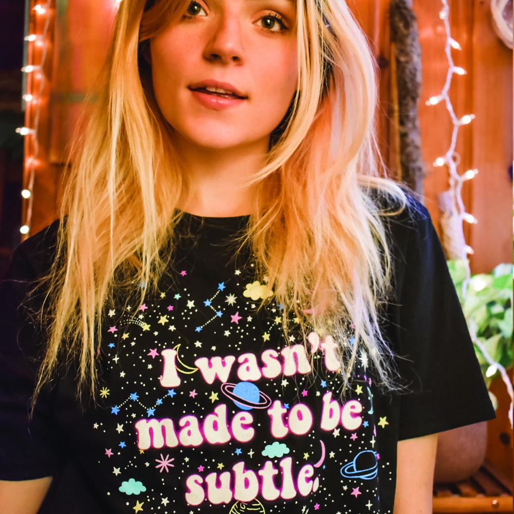 I Wasn’t Made to Be Subtle black graphic t-shirt with colorful space-themed design featuring planets, stars, and moons, modeled by a person with blonde hair – bold cosmic statement tee – The Funnel Cake Tree.