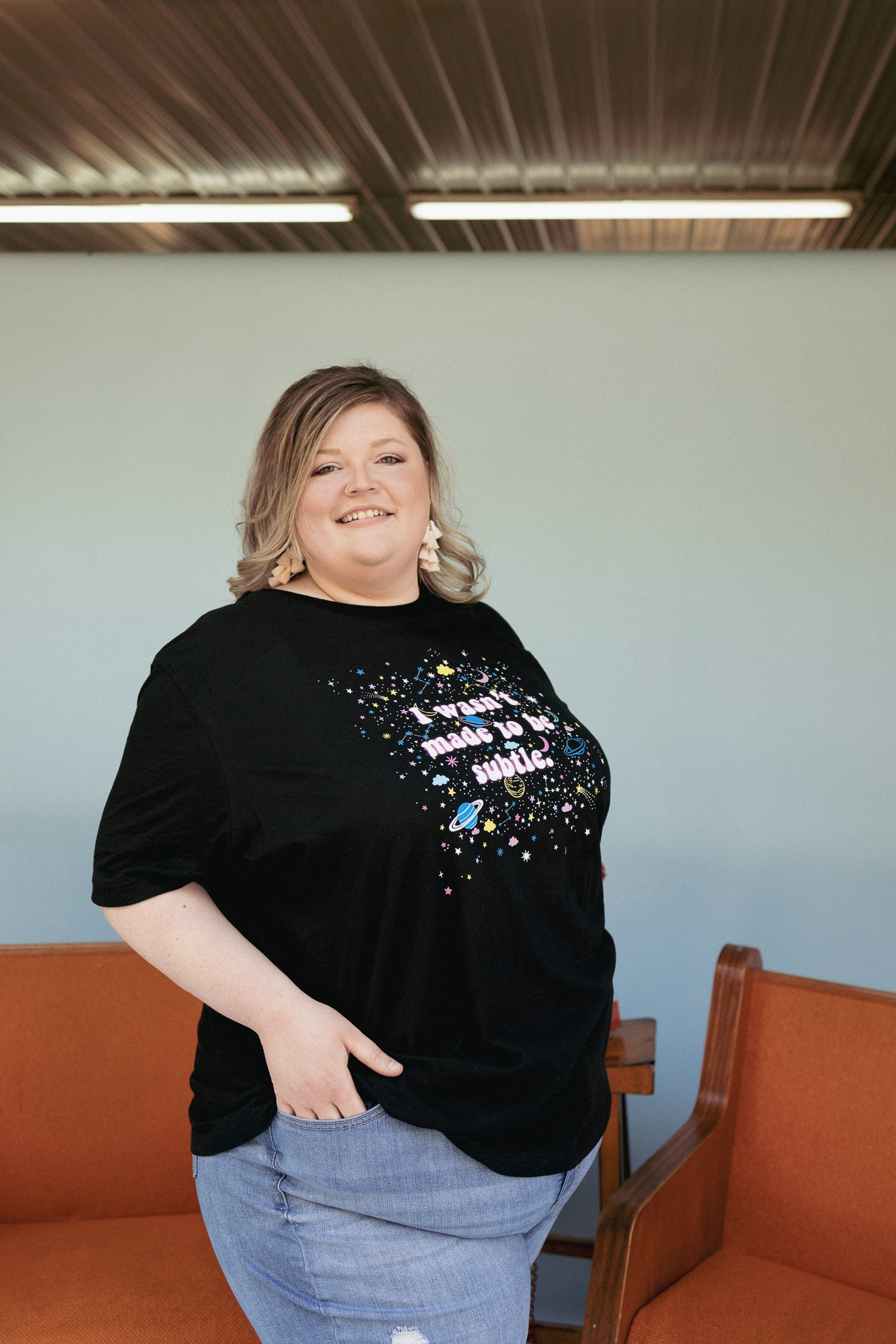 I Wasn’t Made to Be Subtle black graphic t-shirt with colorful planets, stars, and retro space design, modeled by a smiling person wearing light wash jeans – bold cosmic statement tee – The Funnel Cake Tree.