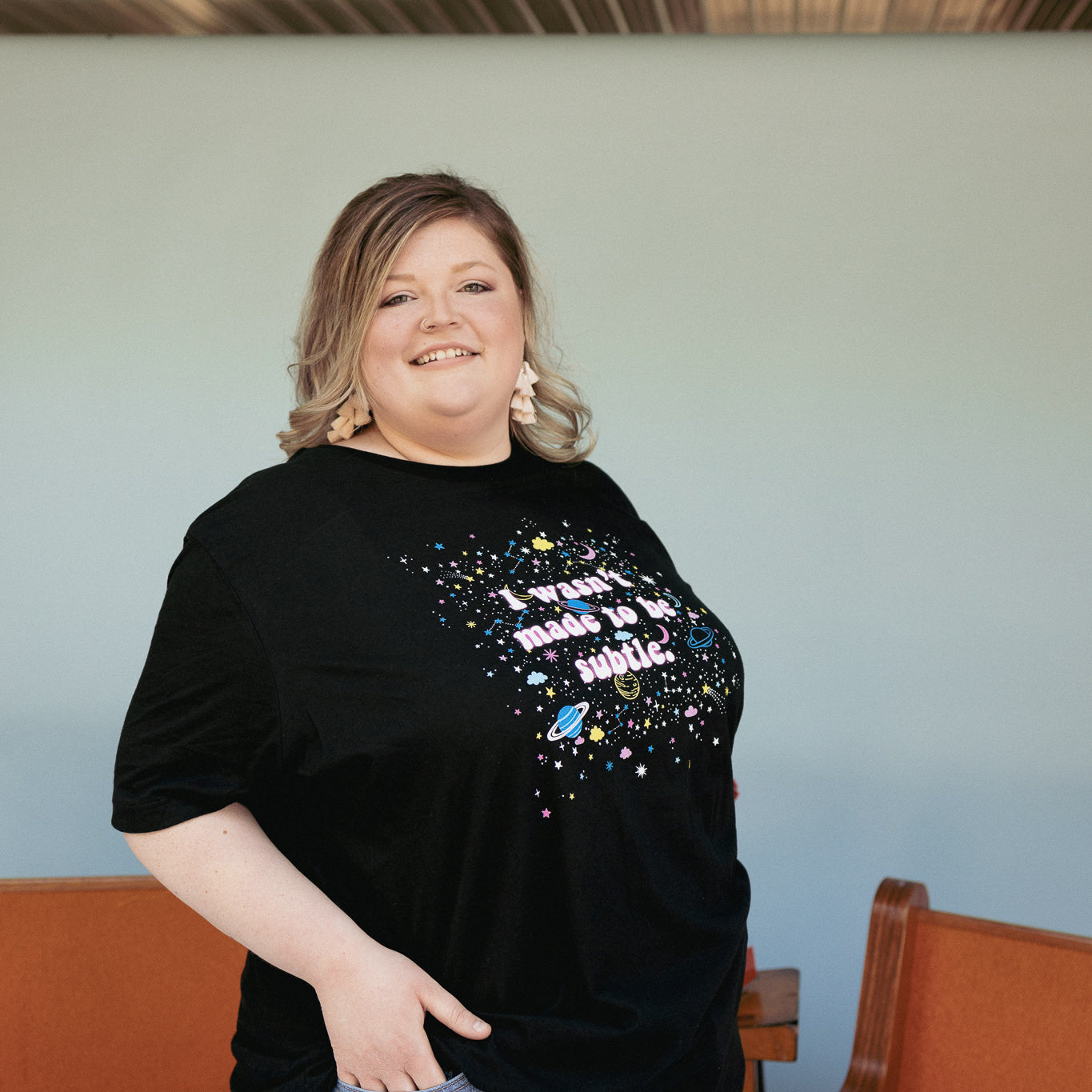 I Wasn’t Made to Be Subtle black graphic t-shirt with colorful planets, stars, and retro space design, modeled by a smiling person wearing light wash jeans – bold cosmic statement tee – The Funnel Cake Tree.