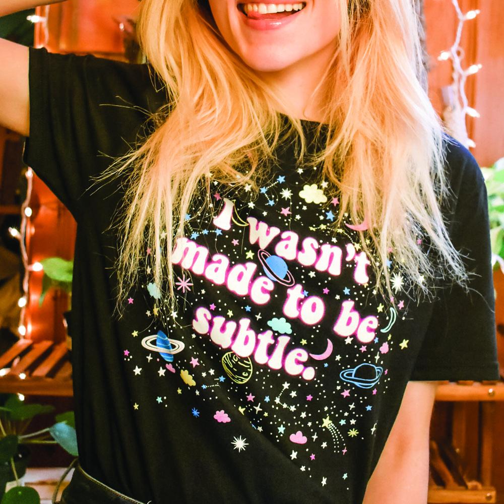 I Wasn’t Made to Be Subtle black graphic t-shirt with colorful stars, planets, and space-themed design, modeled by a person making a peace sign – bold retro cosmic statement tee – The Funnel Cake Tree.