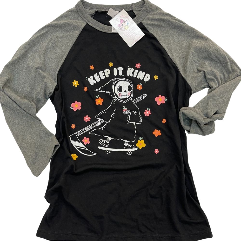 Flat lay of black and grey raglan tee with “Keep It Kind” graphic featuring a cute cartoon grim reaper on a skateboard surrounded by colorful flowers, cozy unisex kindness tee from The Funnel Cake Tree.