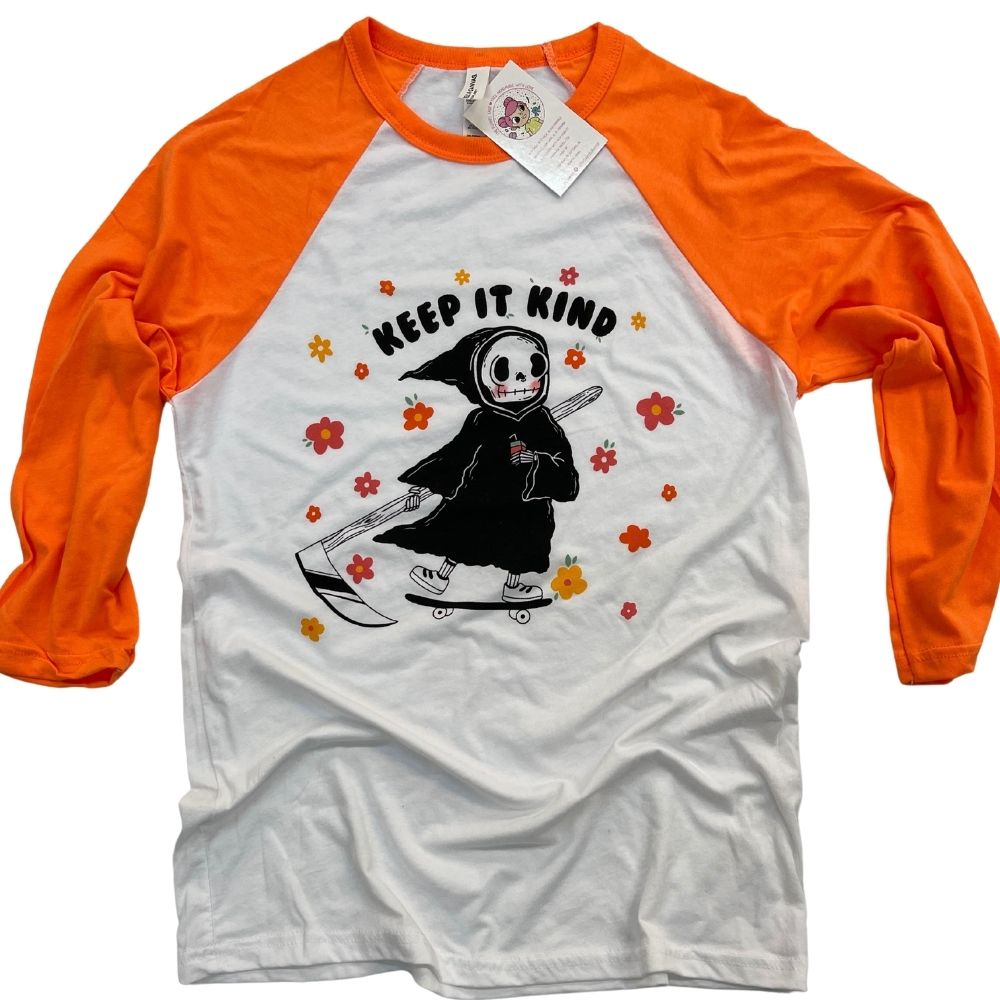 Flat lay of orange and white raglan tee with “Keep It Kind” text and a cute grim reaper holding a coffee and riding a skateboard surrounded by colorful flowers unisex retro kindness graphic tee from The Funnel Cake Tree.
