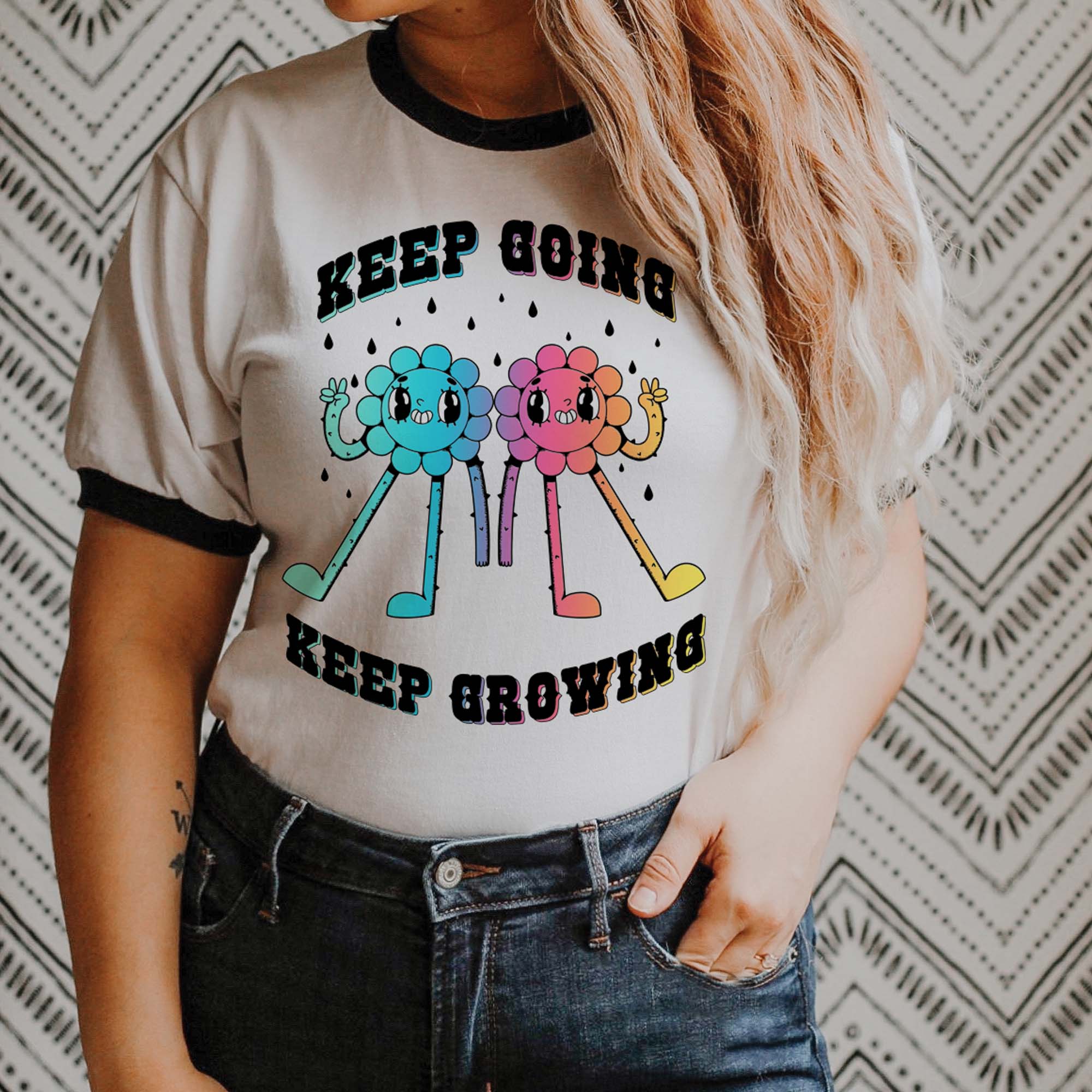Woman wearing a white ringer t-shirt with black trim featuring two colorful cartoon flower characters and the phrase “Keep Going Keep Growing.”