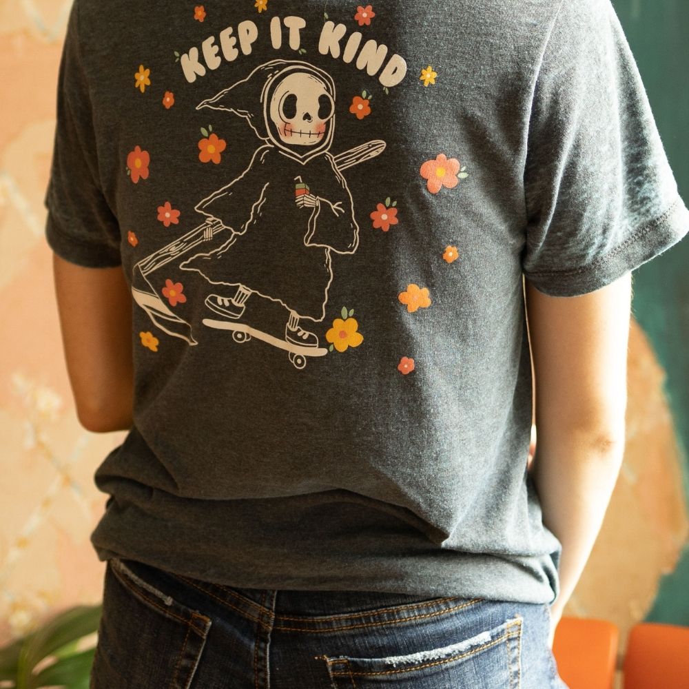 Back view of The Funnel Cake Tree’s charcoal heather graphic tee featuring a cartoon Grim Reaper on a skateboard surrounded by colorful flowers with the text “Keep It Kind.”