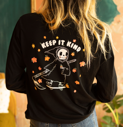 Back view of a woman wearing a black long-sleeve shirt with a large “Keep It Kind” graphic featuring a cute grim reaper riding a skateboard surrounded by flowers