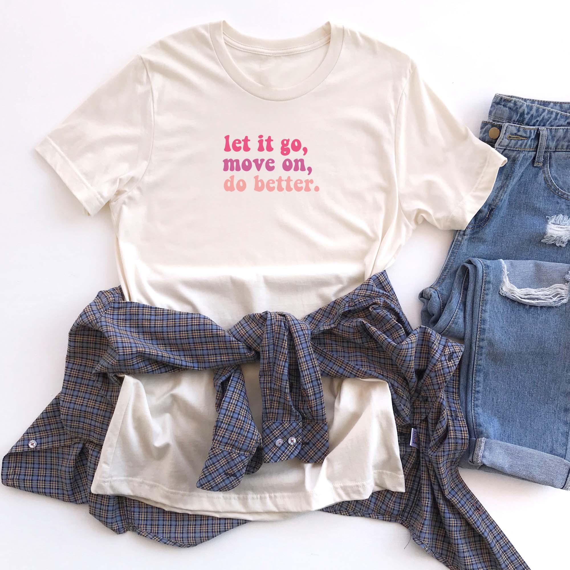 Cream-colored t-shirt with pink and red text that reads “let it go, move on, do better.” displayed with a plaid shirt and denim jeans, The Funnel Cake Tree graphic tee.
