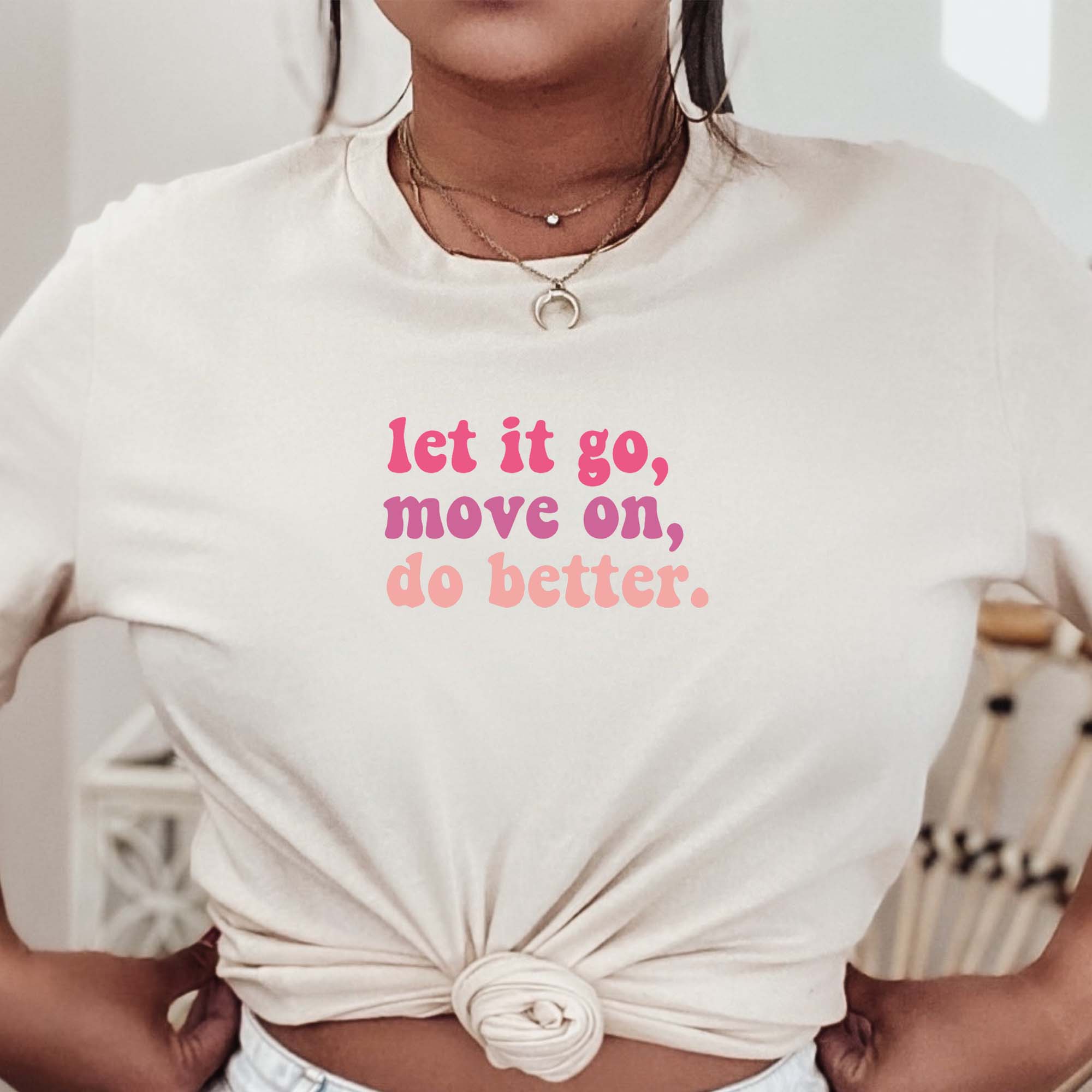 Cream-colored t-shirt with pink and coral text that says “let it go, move on, do better.” worn by a woman styled with knotted front, The Funnel Cake Tree graphic tee.