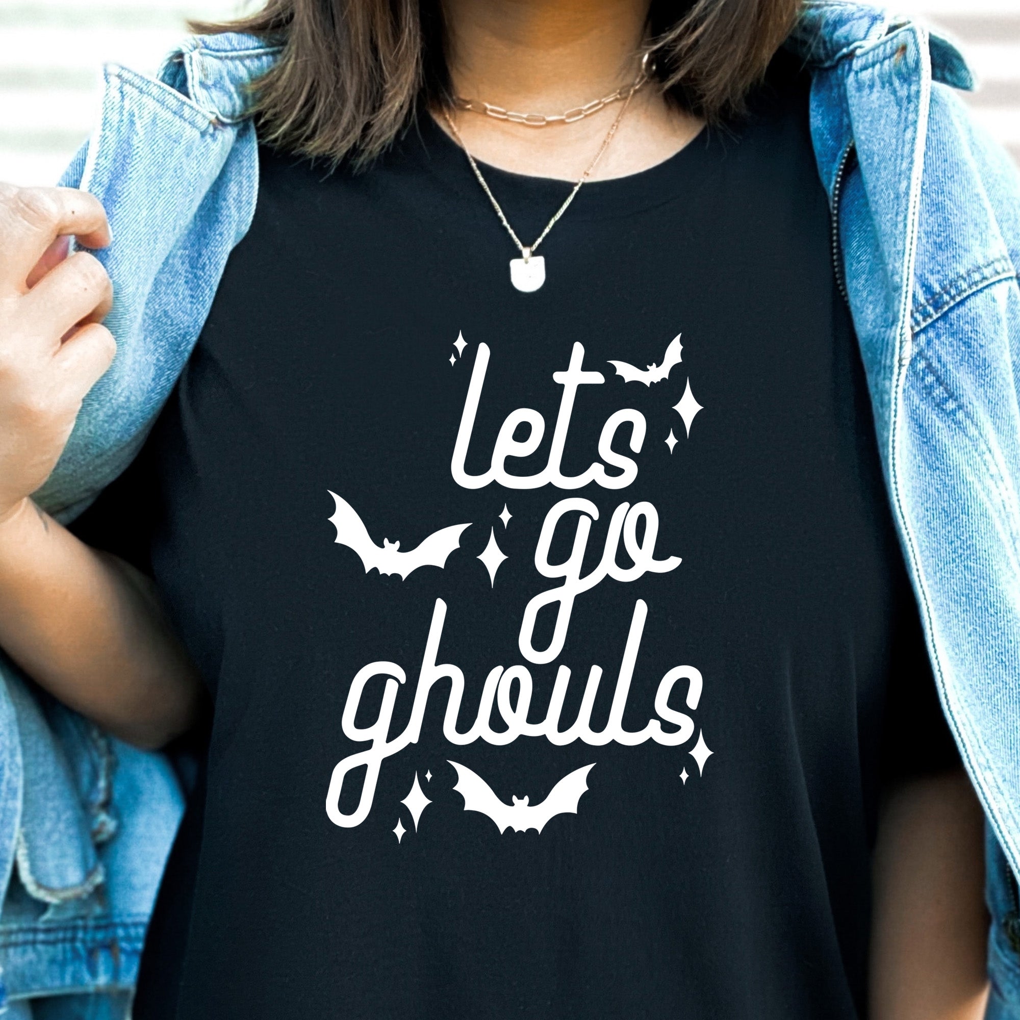 The Funnel Cake Tree “Let’s Go Ghouls” black graphic tee featuring white retro lettering with bats and sparkles, styled with a denim jacket for a fun Halloween-inspired look.