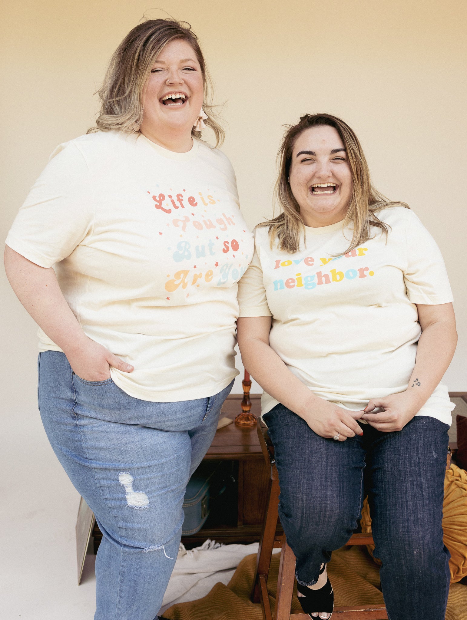 Two smiling people wearing cream-colored graphic t-shirts  one reads “Life is tough but so are you” in colorful retro text with stars, and the other reads “Love your neighbor” in rainbow bubble letters.