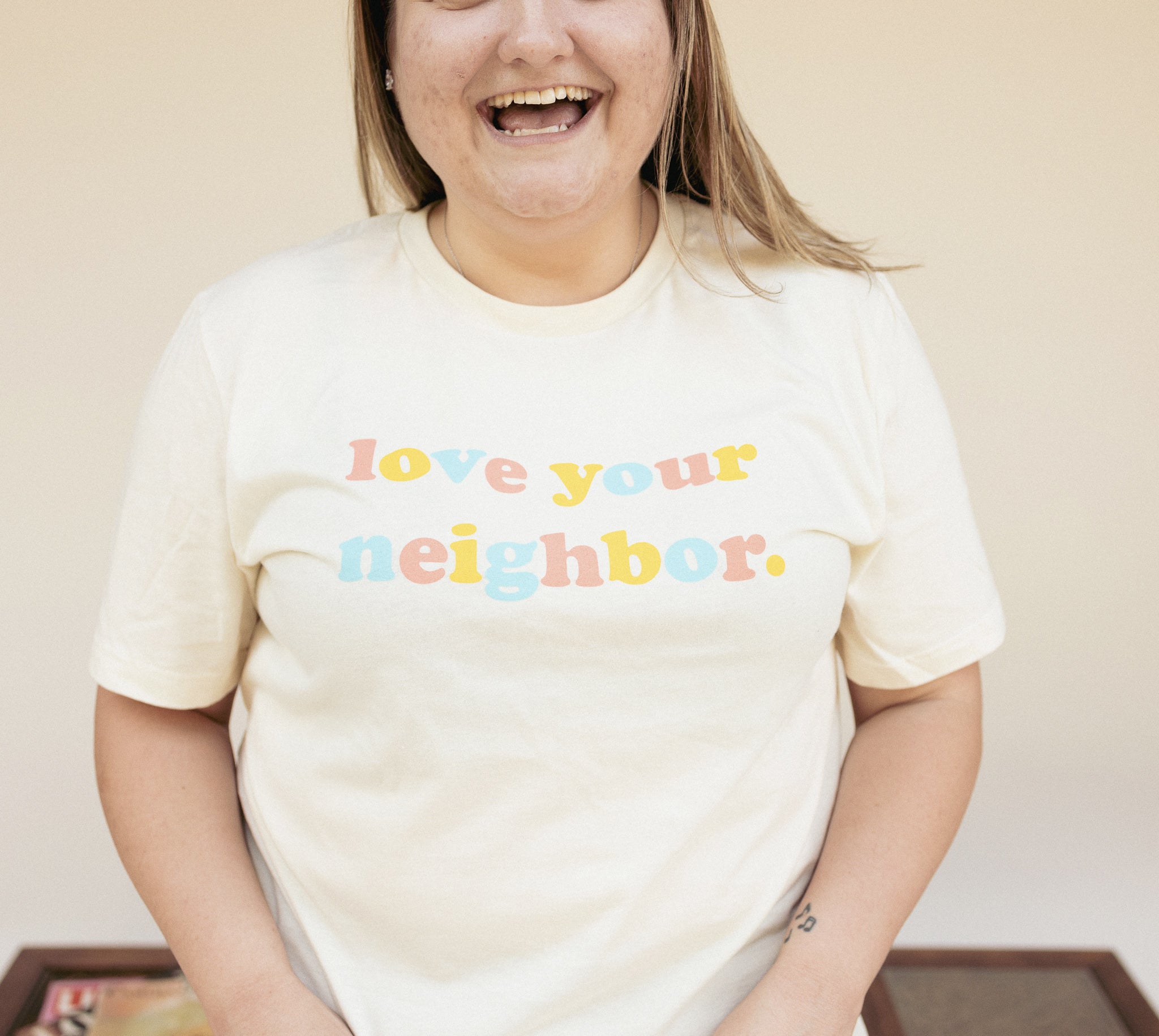 Love Your Neighbor cream graphic t-shirt with colorful retro text in yellow, pink, and blue, modeled by a smiling person. Soft cotton positive message tee – The Funnel Cake Tree.