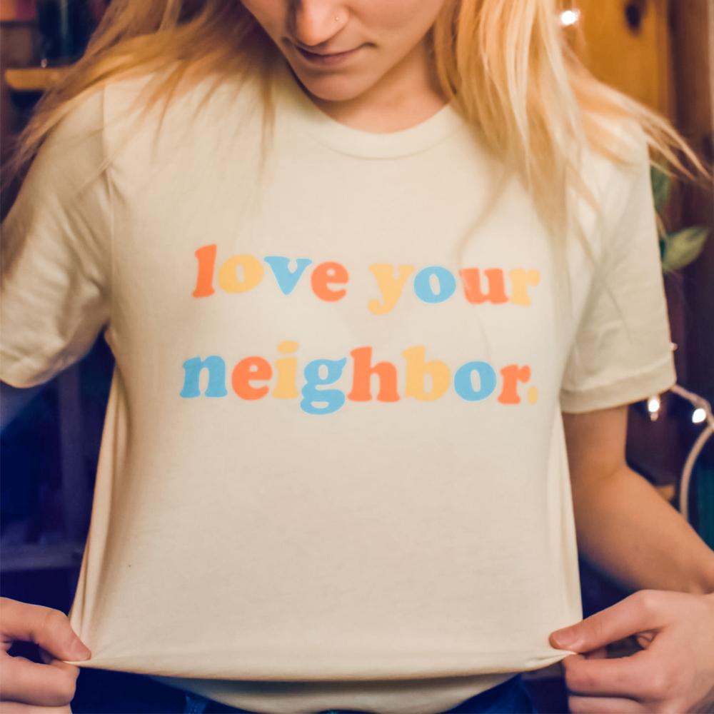 Love Your Neighbor cream graphic t-shirt with bold retro rainbow letters in orange, yellow, and blue, modeled by a person holding the bottom of the shirt – uplifting positive message tee – The Funnel Cake Tree.