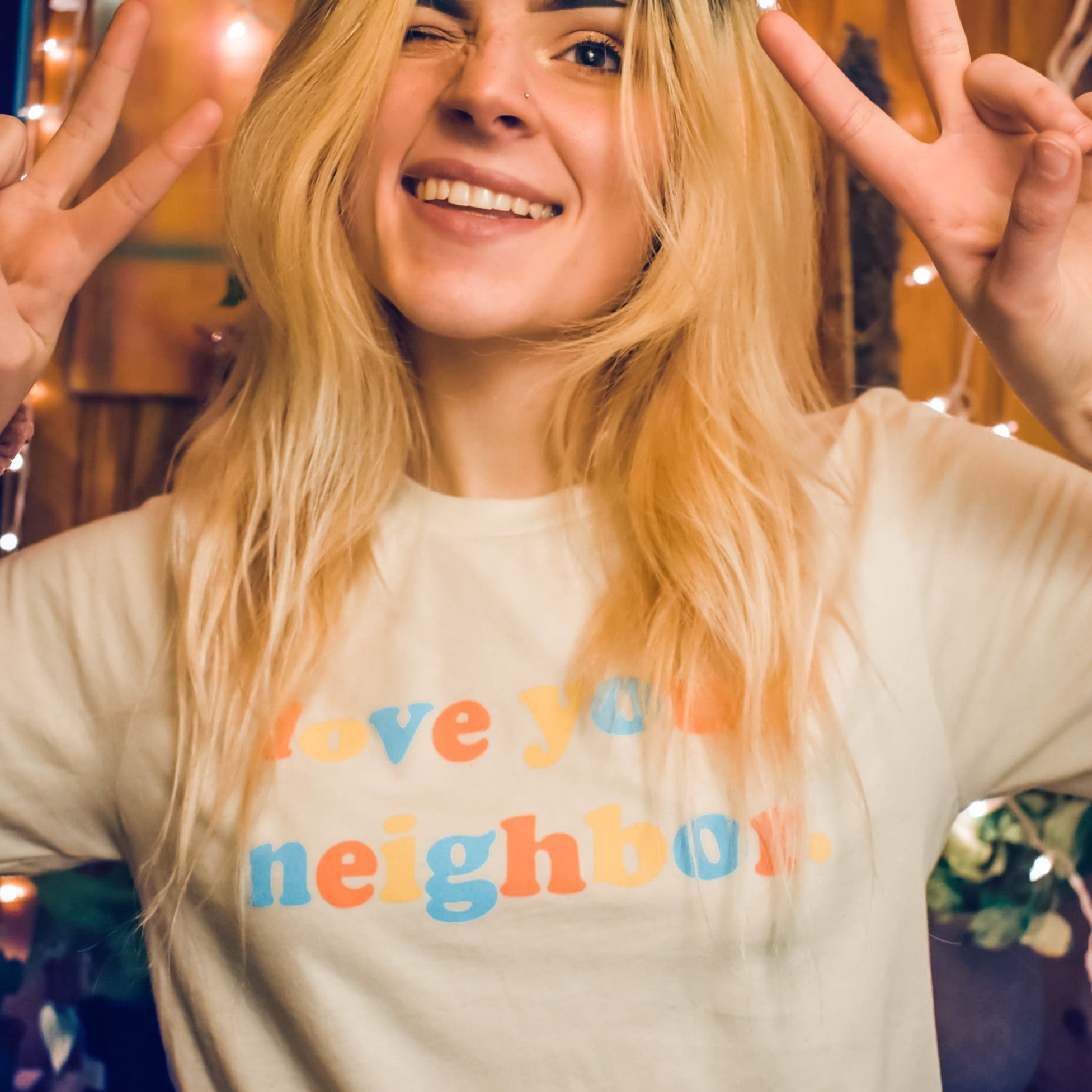 Isabel from the funnel cake tree smiling holding both hands in a peace sign wearing a natural colored tshirt the words love your neighbor printed on the front.