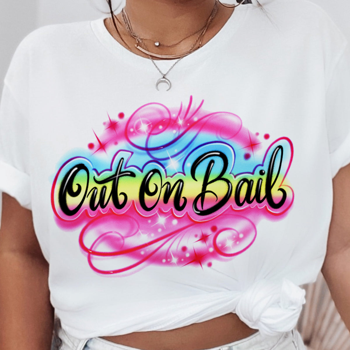 Model wearing a white t-shirt with a vibrant pink, blue, and yellow Out On Bail airbrush-style graphic, styled with light-wash denim — The Funnel Cake Tree retro graphic tee.