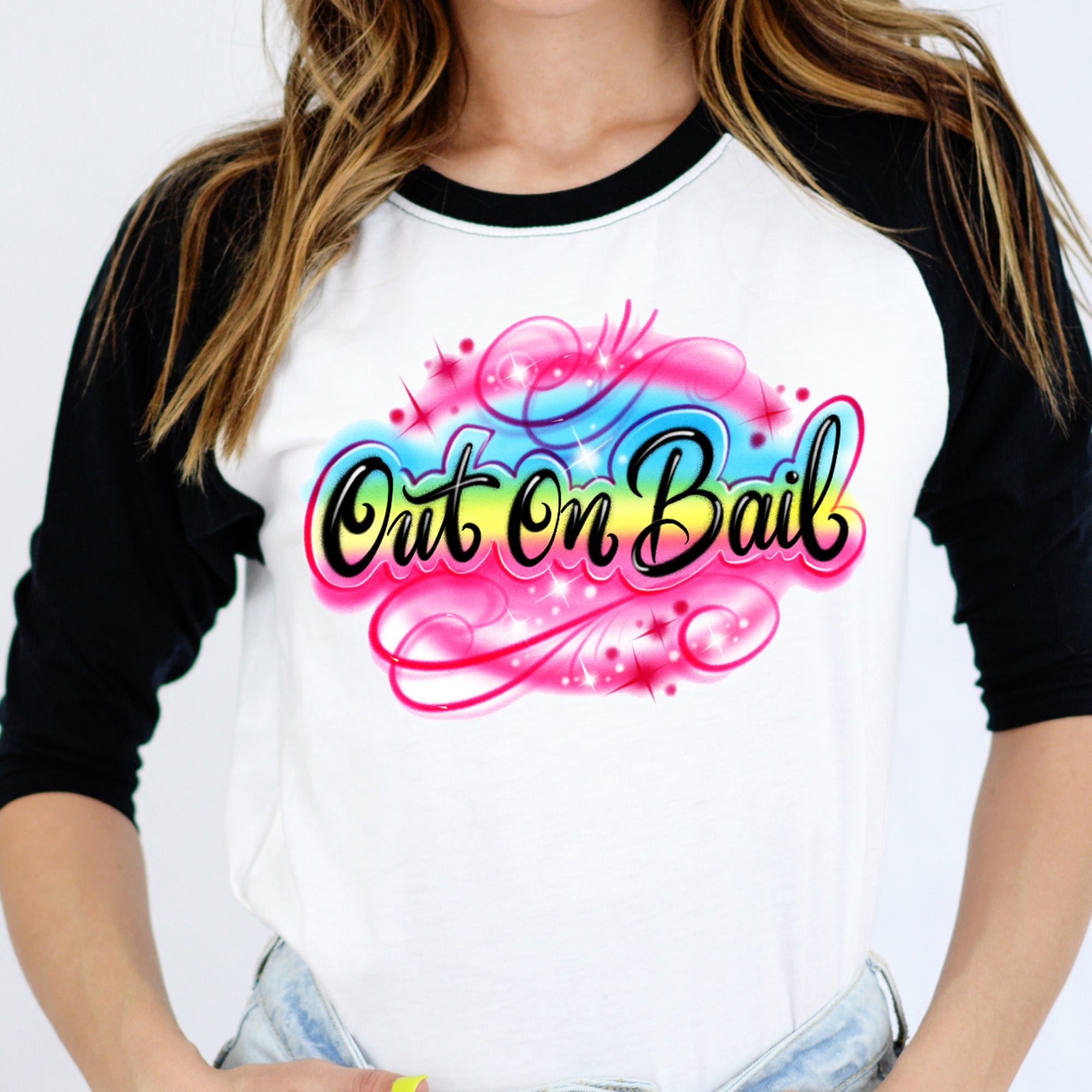 Model wearing a white raglan tee with black sleeves featuring a vibrant pink, blue, and yellow Out On Bail airbrush-style graphic — The Funnel Cake Tree retro graphic tee.