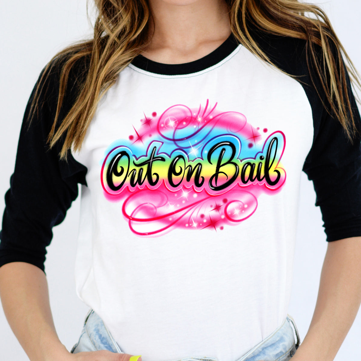 Model wearing a white raglan tee with black sleeves featuring a vibrant pink, blue, and yellow Out On Bail airbrush-style graphic — The Funnel Cake Tree retro graphic tee.