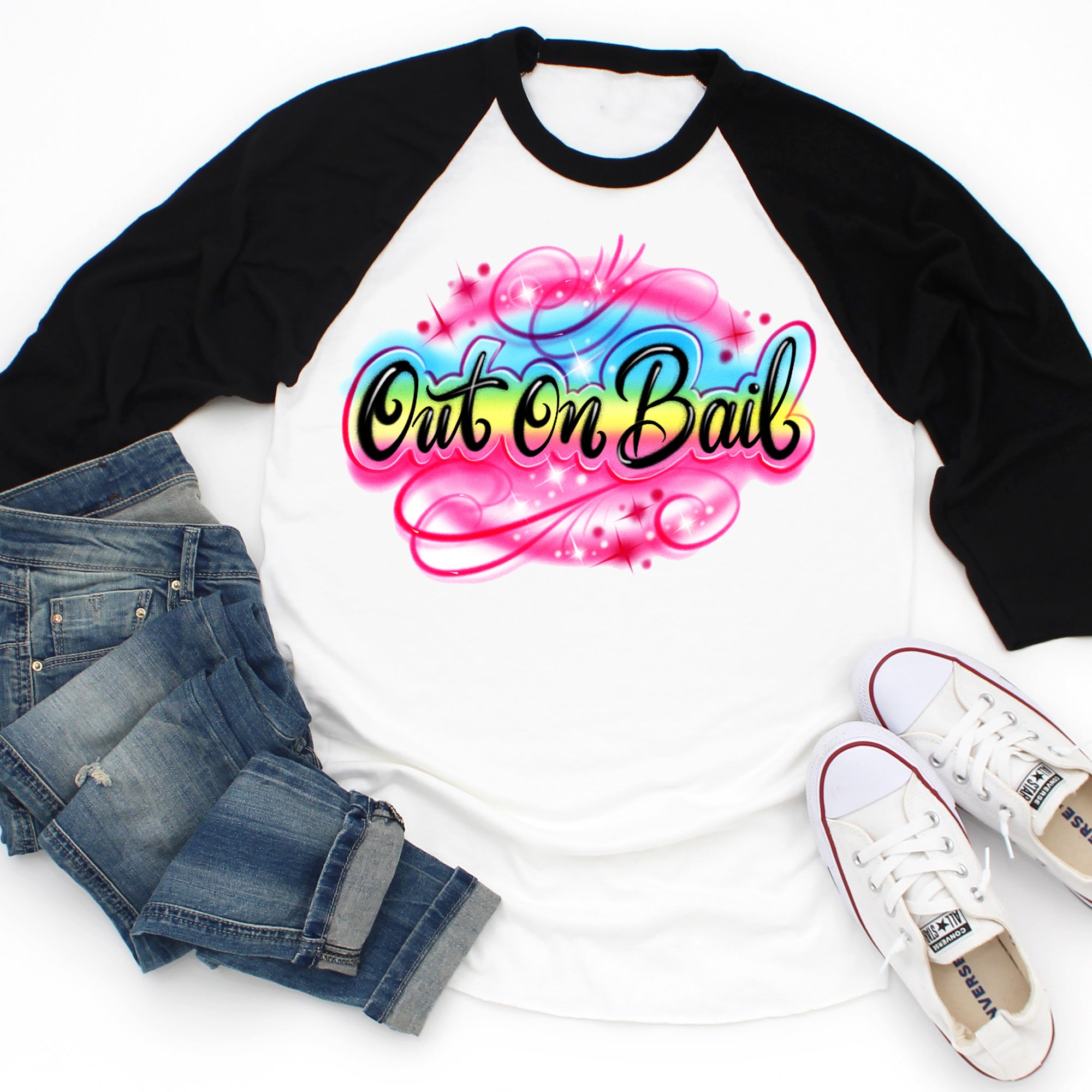 Flat lay of a white raglan tee with black sleeves featuring a colorful “Out On Bail” airbrush-style graphic, styled with jeans and white sneakers — The Funnel Cake Tree.