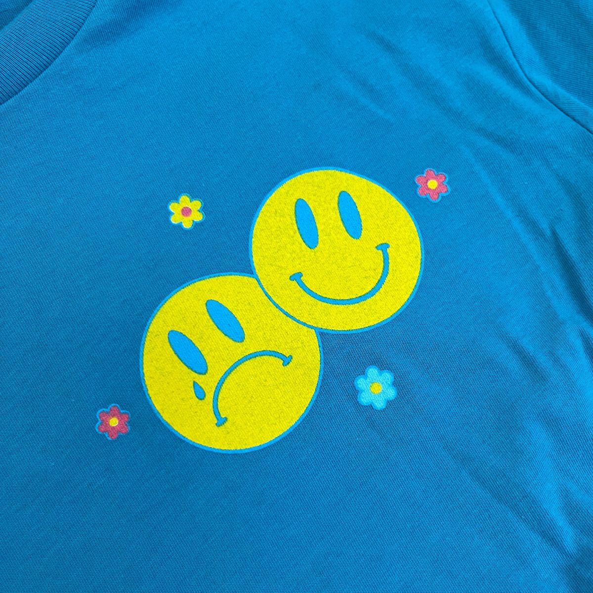 Blue graphic t-shirt with two yellow smiley faces and colorful retro flower accents – Sometimes Sad Always Rad design – The Funnel Cake Tree.