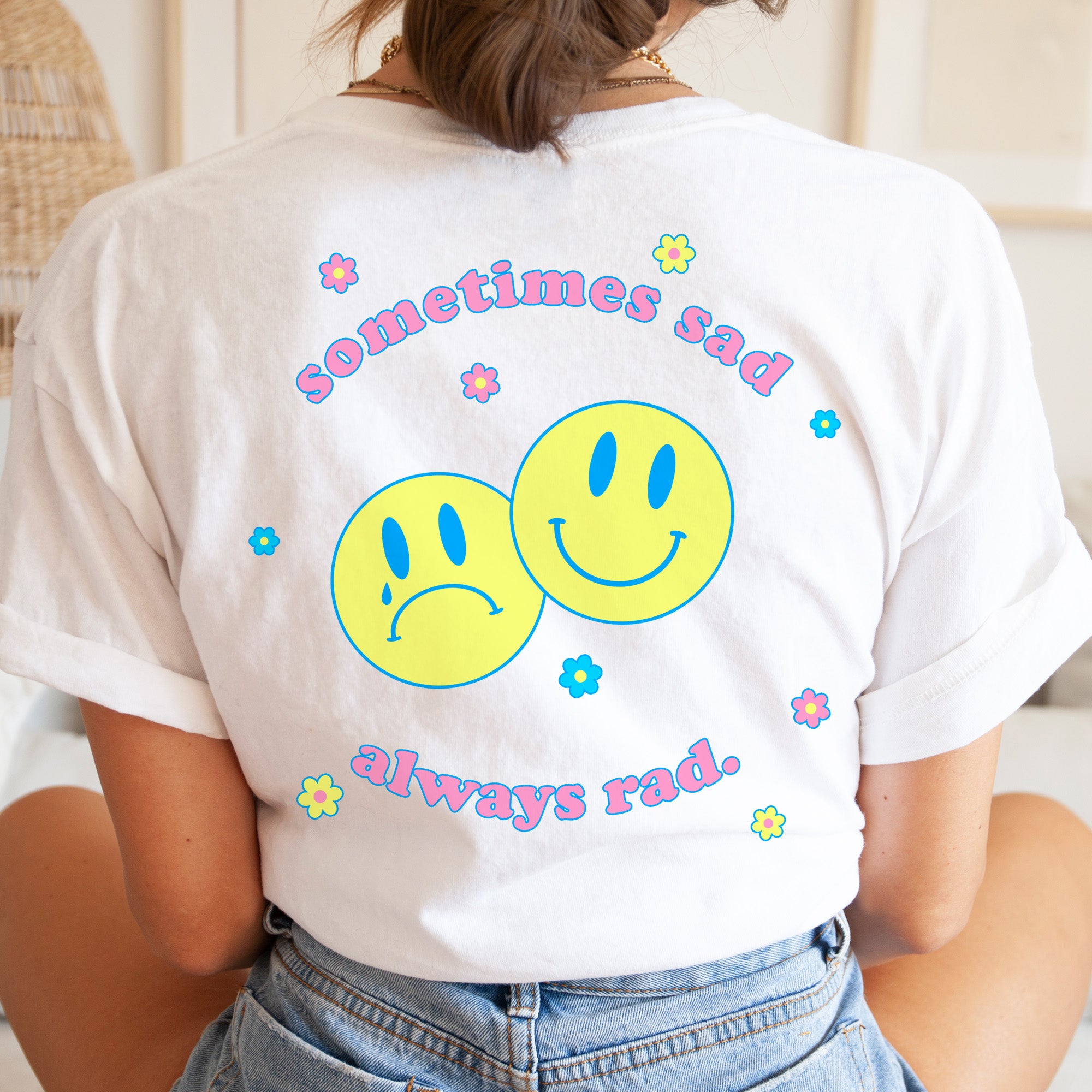 White graphic t-shirt with colorful retro design that says “Sometimes Sad Always Rad” with two yellow smiley faces and pastel flowers – The Funnel Cake Tree.
