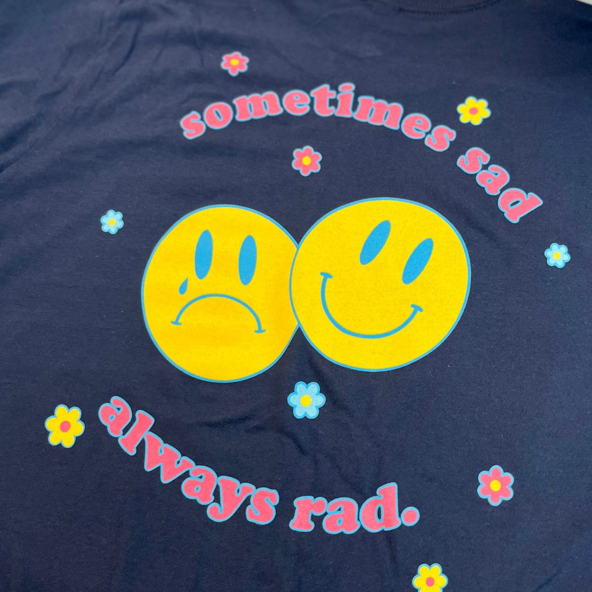 Navy blue graphic t-shirt with bright yellow smiley faces and pastel flowers, featuring the text “Sometimes Sad Always Rad” in retro pink font – The Funnel Cake Tree.