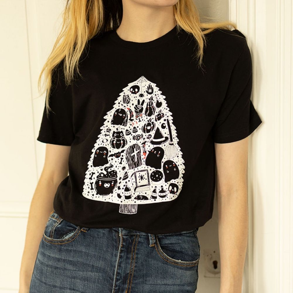 Model wearing The Funnel Cake Tree black graphic tee featuring a spooky Christmas tree made up of cute and creepy Halloween icons like ghosts, coffins, skulls, cauldrons, and potions printed in white and red.