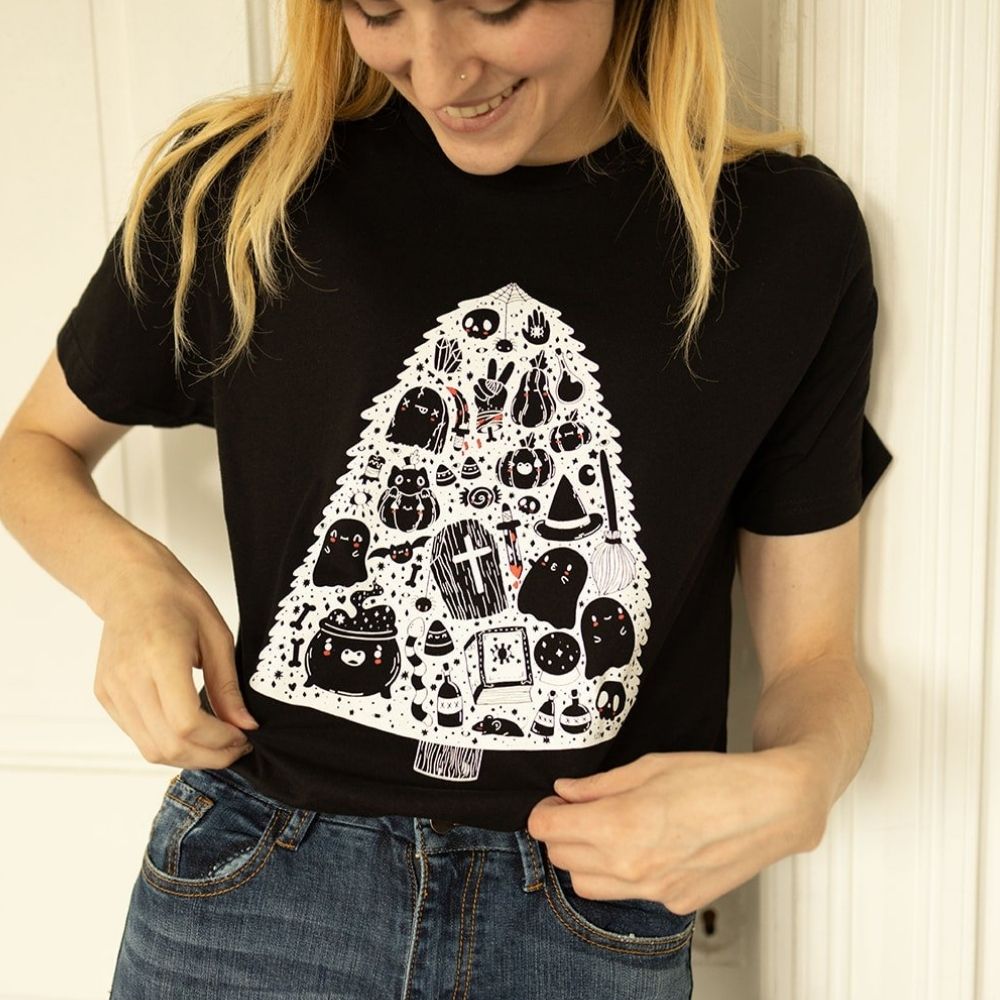 Woman wearing The Funnel Cake Tree black graphic t-shirt featuring a spooky tree design made of ghosts, skulls, bats, potions, and other Halloween icons printed in white and red.