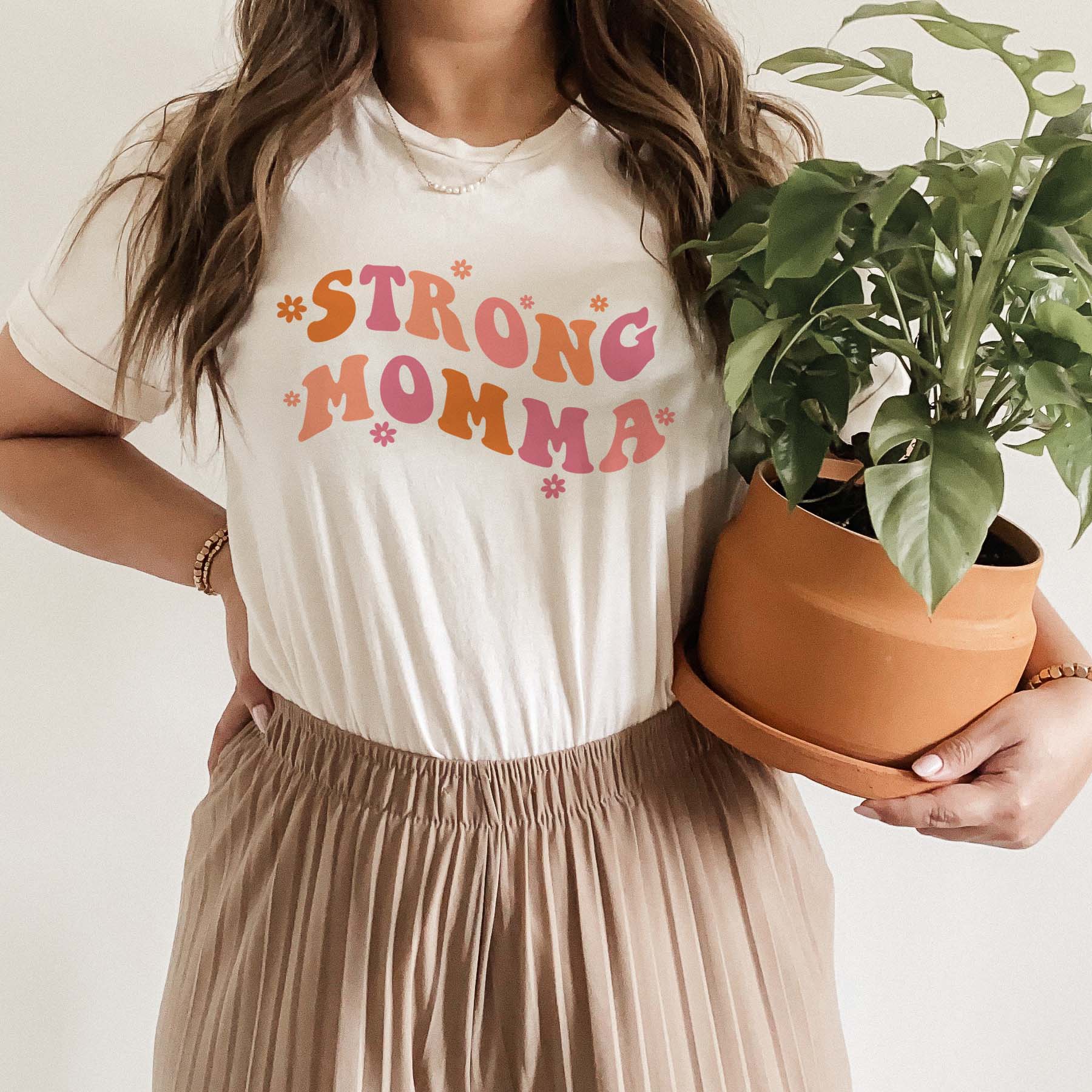 Woman wearing a cream-colored graphic tee with the words “Strong Momma” in retro pink and orange letters, holding a potted plant.