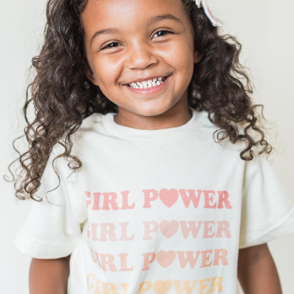 Smiling girl wearing a white fabric t-shirt with Girl Power printed in a repeating pastel rainbow pattern.