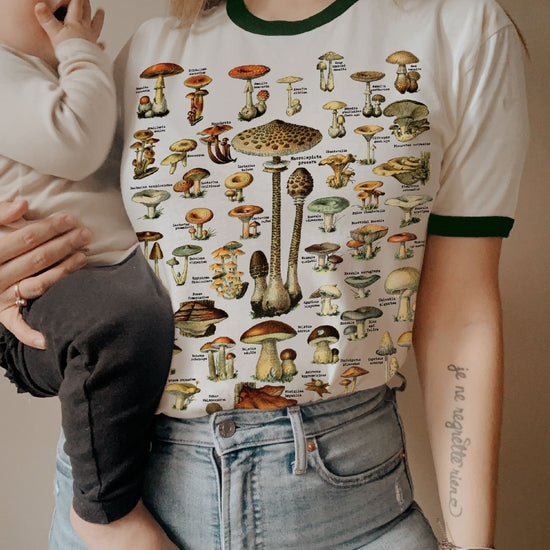 Person wearing a cream ringer t-shirt with green trim featuring a detailed vintage mushroom chart illustration from The Funnel Cake Tree, styled with high-waisted jeans and holding a baby.