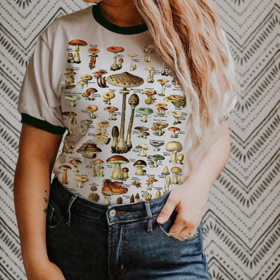 Person wearing a cream ringer t-shirt with green trim featuring a vintage mushroom illustration chart, styled with high-waisted denim — The Funnel Cake Tree graphic tee.