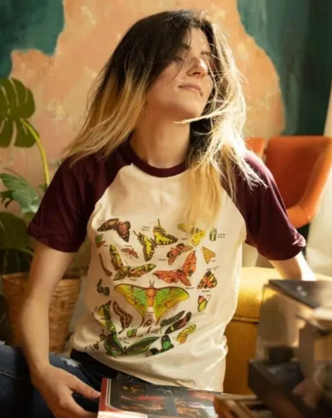 Woman wearing a cream and burgundy raglan tee featuring a vintage butterfly and moth illustration by The Funnel Cake Tree, sitting in a warmly lit room.