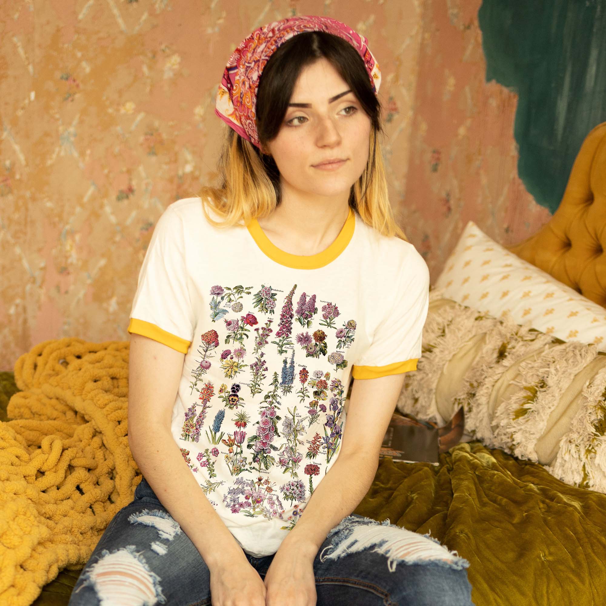 Woman wearing a cream ringer t-shirt with yellow trim featuring a colorful vintage wildflower illustration chart, sitting on a cozy bed.