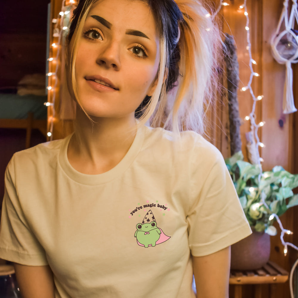 Model wearing The Funnel Cake Tree “You’re Magic Baby” cream graphic tee featuring a cute green frog in a wizard hat and pink cape with magical star accents.