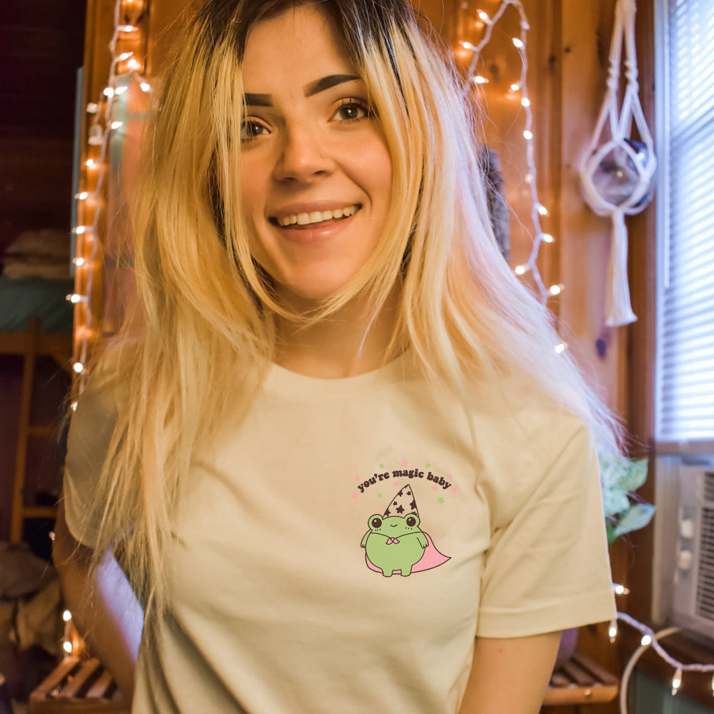 Smiling person wearing a beige graphic t-shirt with a cute green wizard frog, pink cape, and text “You’re magic baby”. The Funnel Cake Tree.