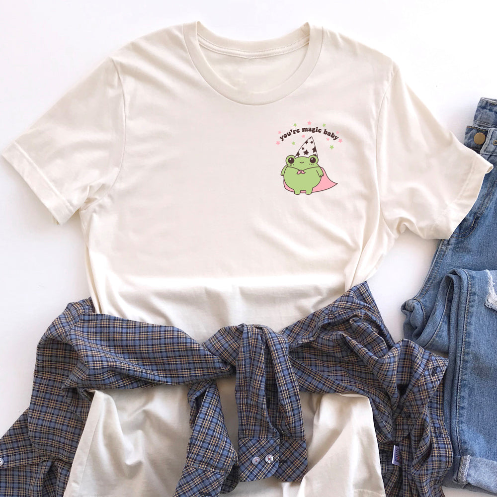 Flat lay of a cream-colored graphic t-shirt featuring a cute green frog in a wizard hat and pink cape with the phrase “You’re magic baby,” styled with plaid shirt and jeans – The Funnel Cake Tree.