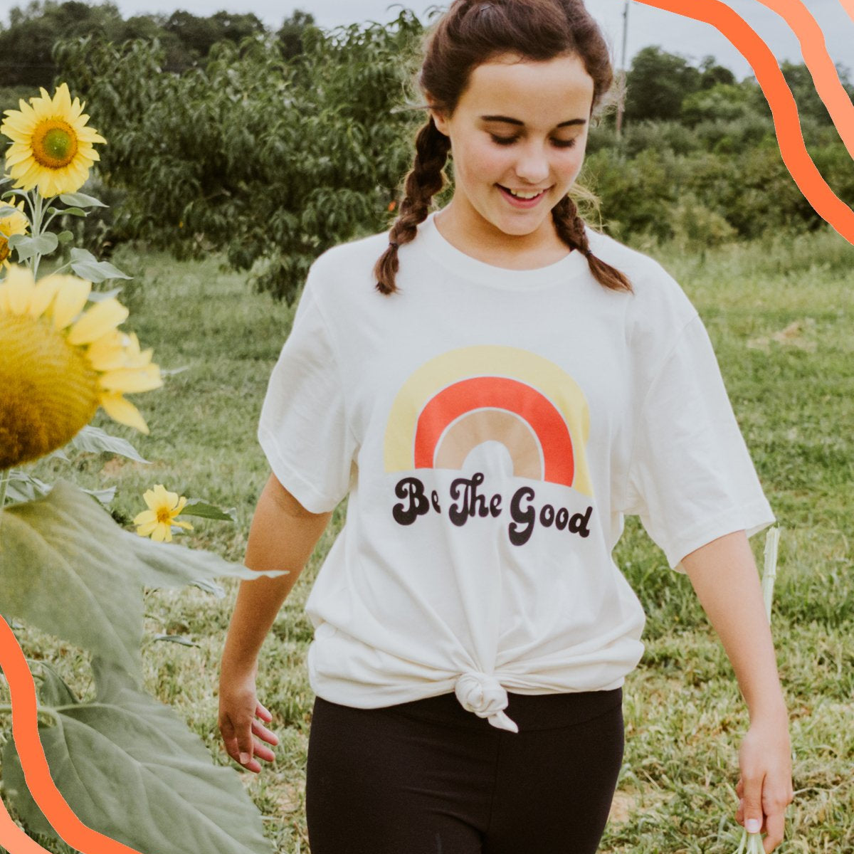Woman walking through a sunflower field wearing a white ‘Be The Good’ t-shirt with a retro rainbow design and smiling in a natural outdoor setting.