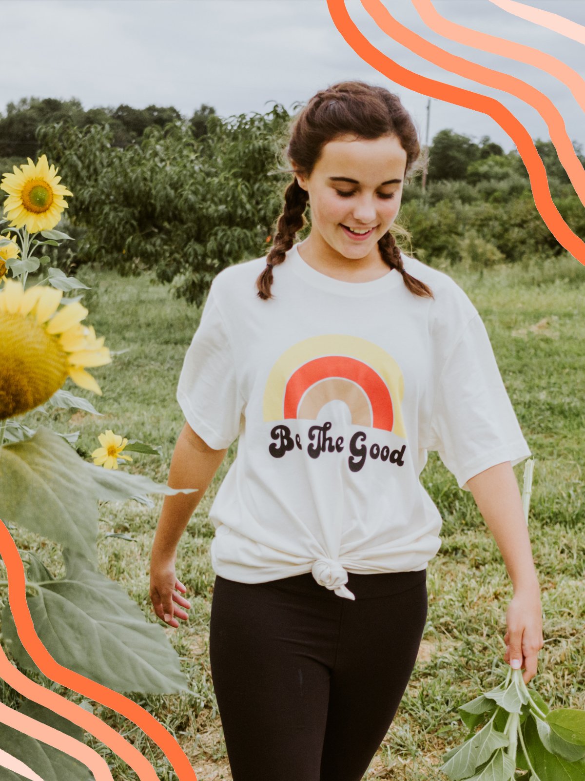 Woman walking through a sunflower field wearing a white ‘Be The Good’ t-shirt with a retro rainbow design and smiling in a natural outdoor setting.