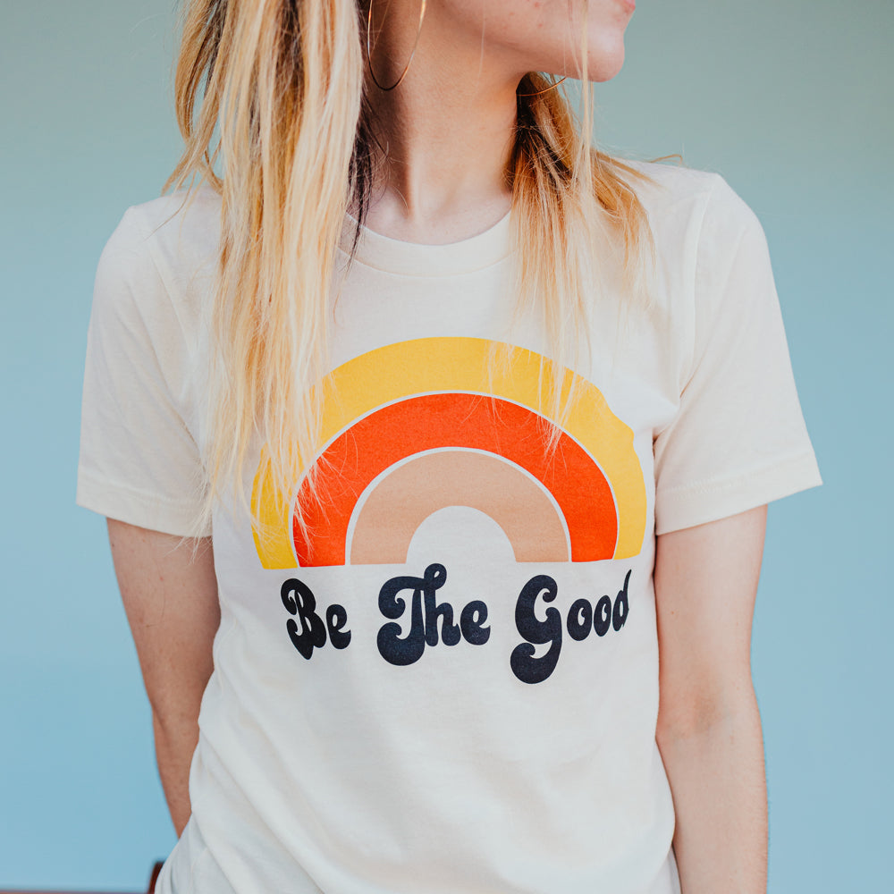 Close-up of a woman wearing a cream ‘Be The Good’ t-shirt with a retro rainbow design in orange, yellow, and tan tones against a blue background.