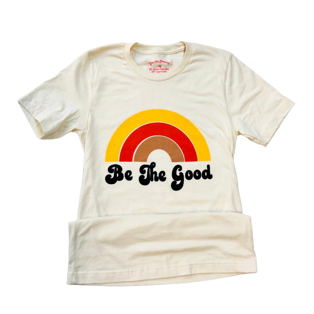 Flat lay of a cream ‘Be The Good’ t-shirt with a retro rainbow design in red, yellow, and tan tones on a white background.