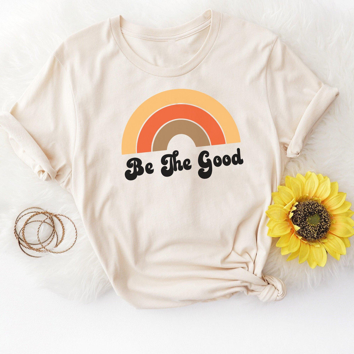 Beige t-shirt with retro rainbow design and bold black text reading ‘Be The Good,’ styled with a sunflower and gold bracelets on a soft white background.