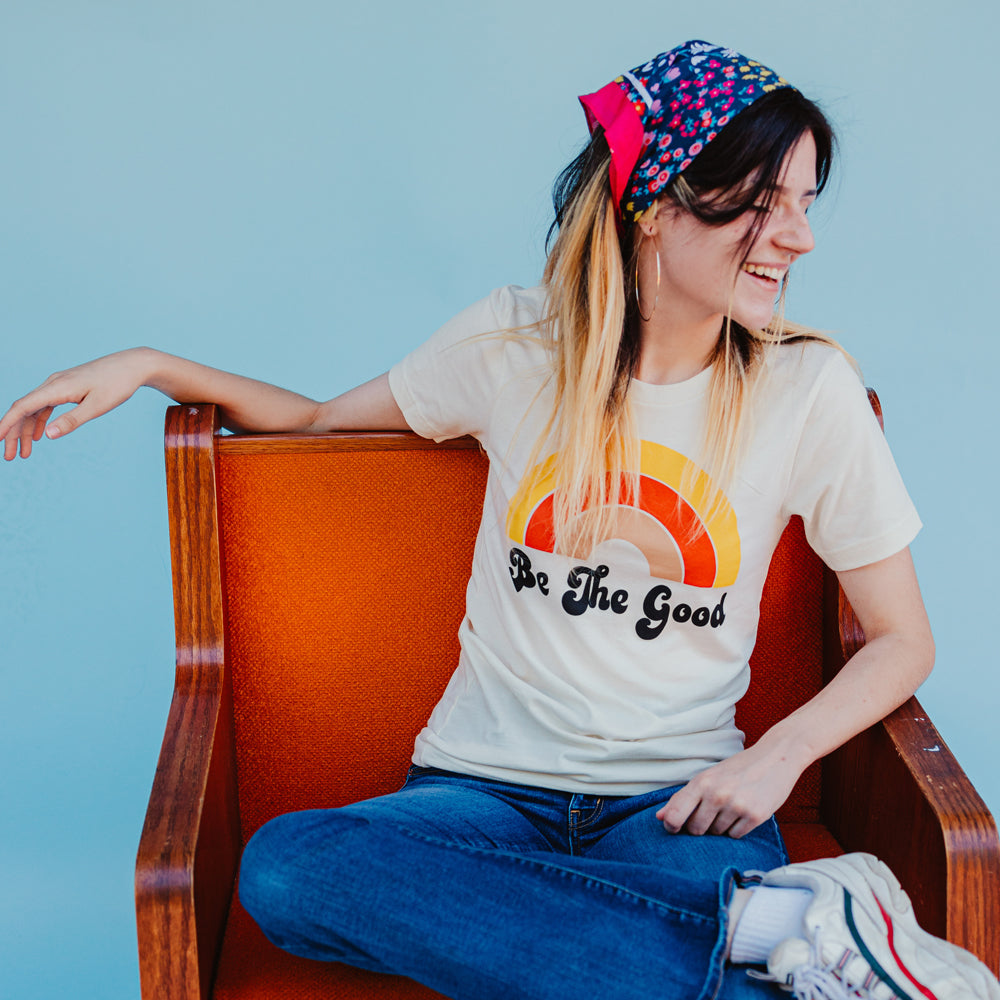 Woman sitting on an orange chair wearing a cream ‘Be The Good’ t-shirt with a retro rainbow design, smiling against a light blue background.