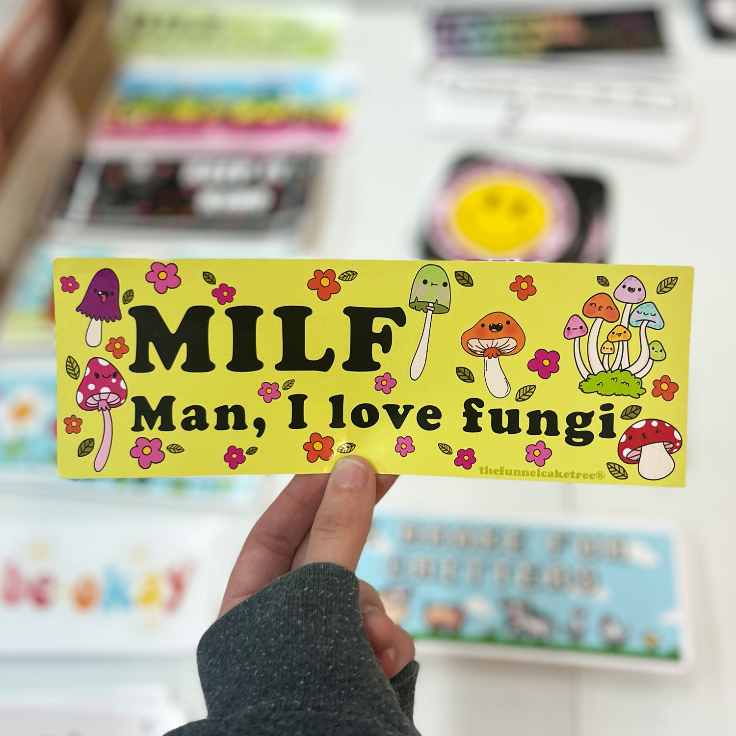 A person holding a yellow bumper sticker with the text 'MILF Man, I love fungi' and illustrated mushrooms.