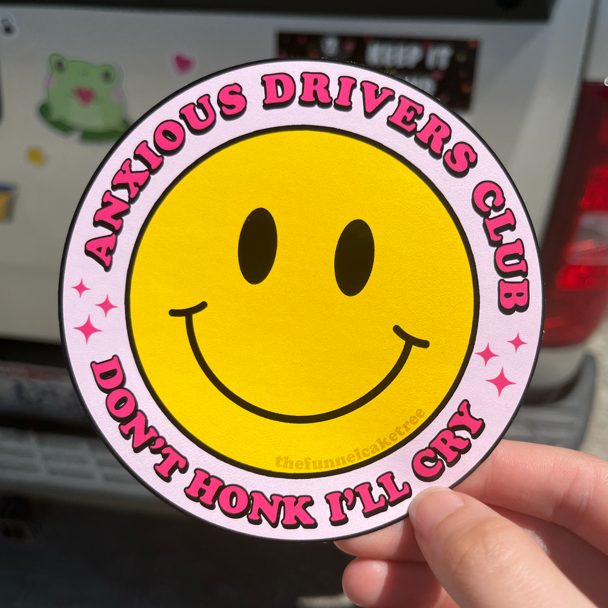 A round yellow bumper sticker with a smiley face and the text 'Anxious Drivers Club' and 'DON'T HONK I'LL CRY' on it, held up against the rear window of a vehicle.
