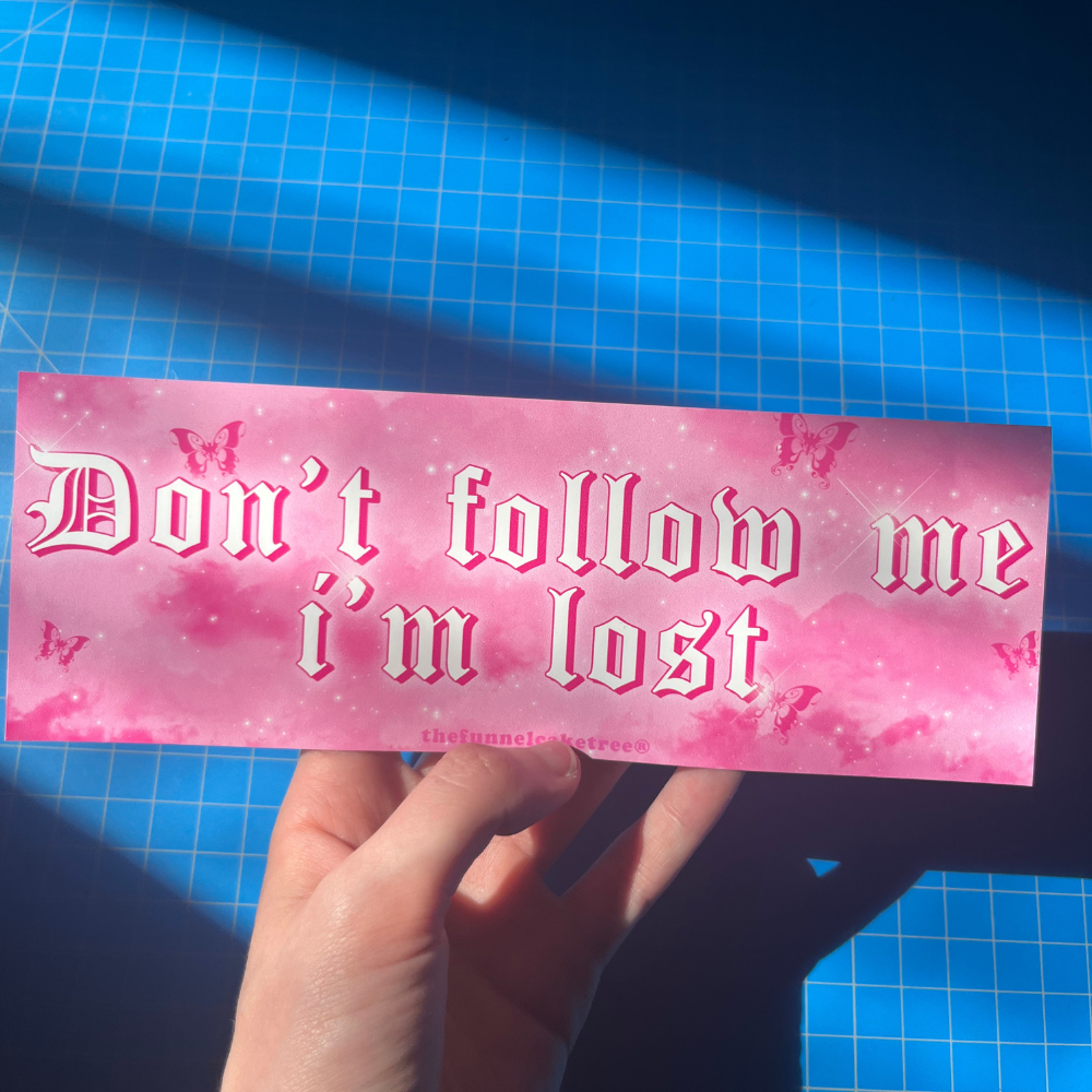 A hand holding a pink sticker with the text 'Don't follow me I'm lost' on it, placed against a blue grid background.