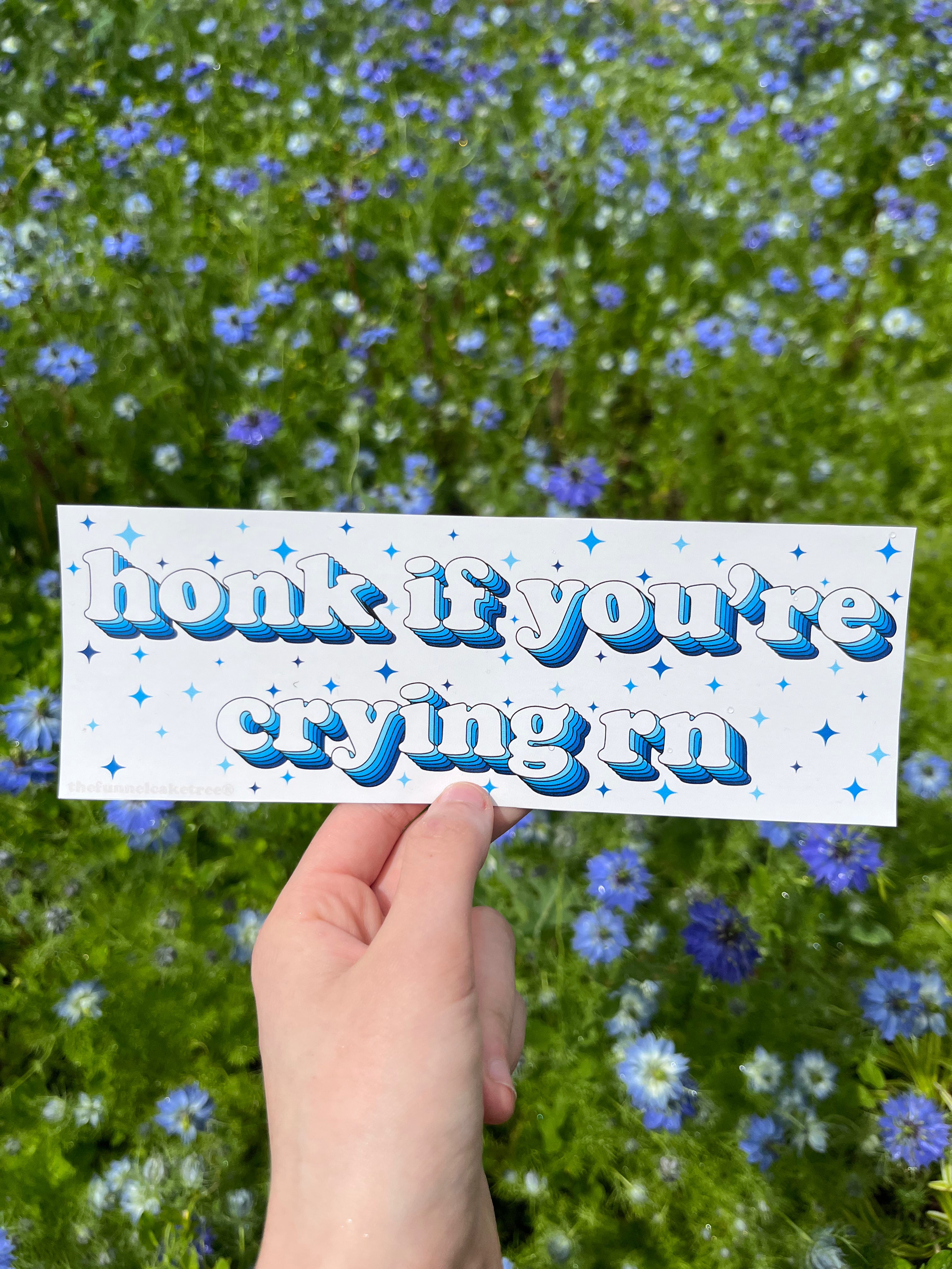 A hand holding a bumper sticker with the text 'Honk if you're crying 😔' written in blue, against a background of a floral field.