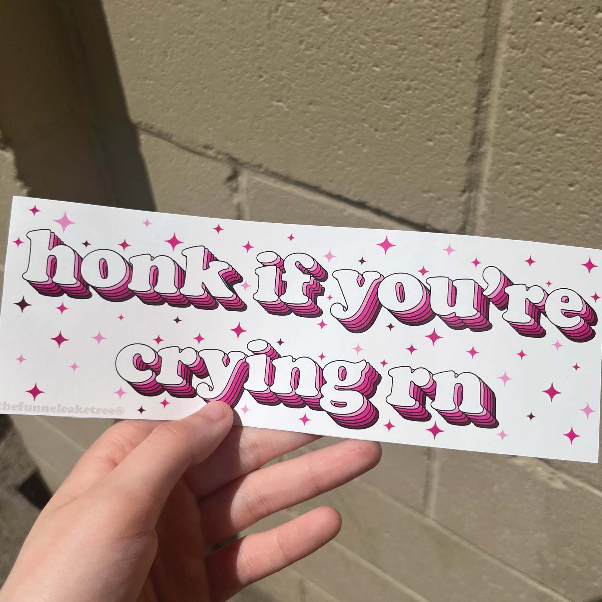Funny vinyl bumper sticker that says ‘Honk if you're crying rn’ in pink font on a light colored background — waterproof and UV-protected, perfect for cars.