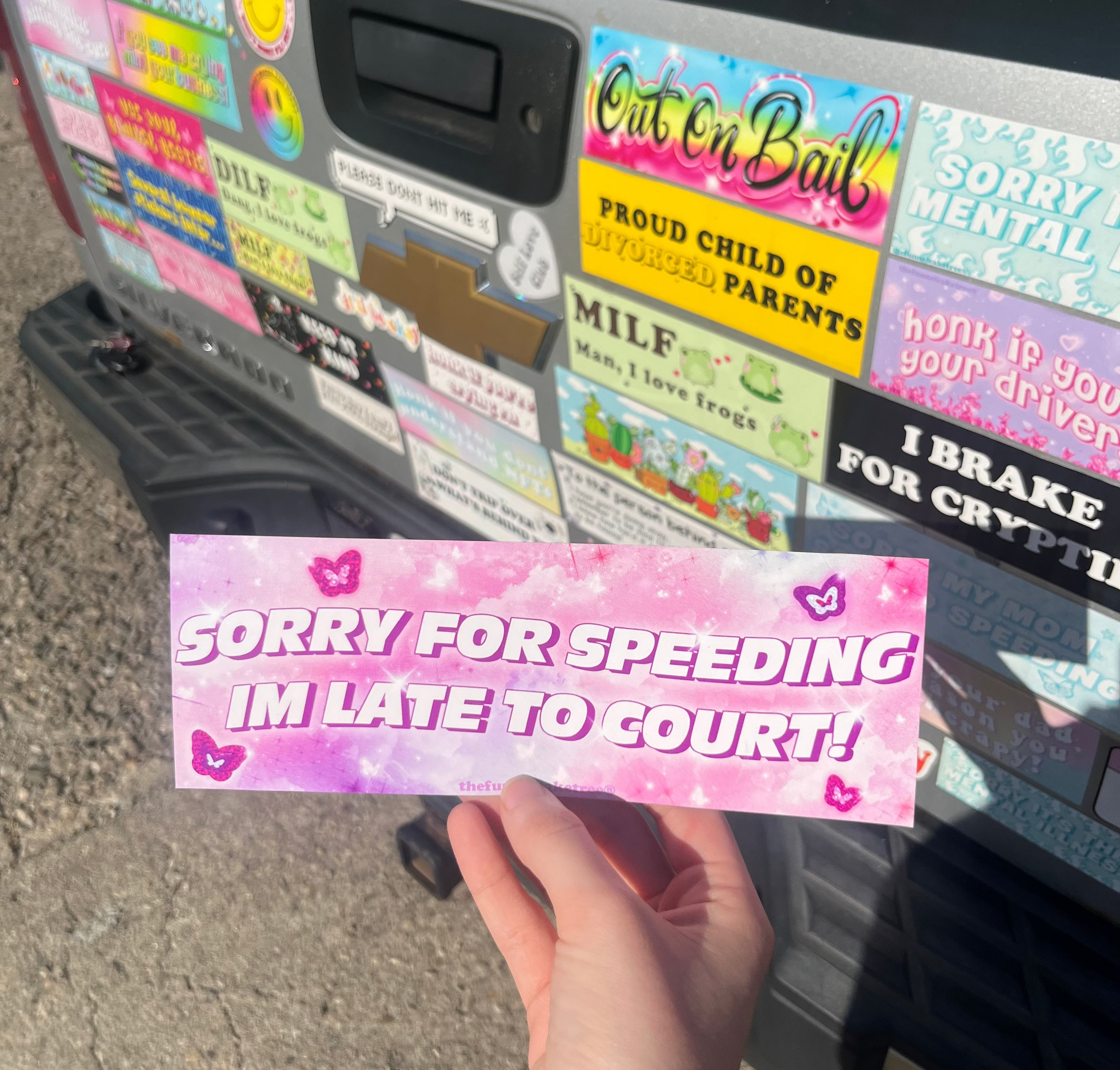 A hand holding a pink bumper sticker with text 'SORRY FOR SPEEDING IM LATE TO COURT' on a background showing other various bumper stickers.