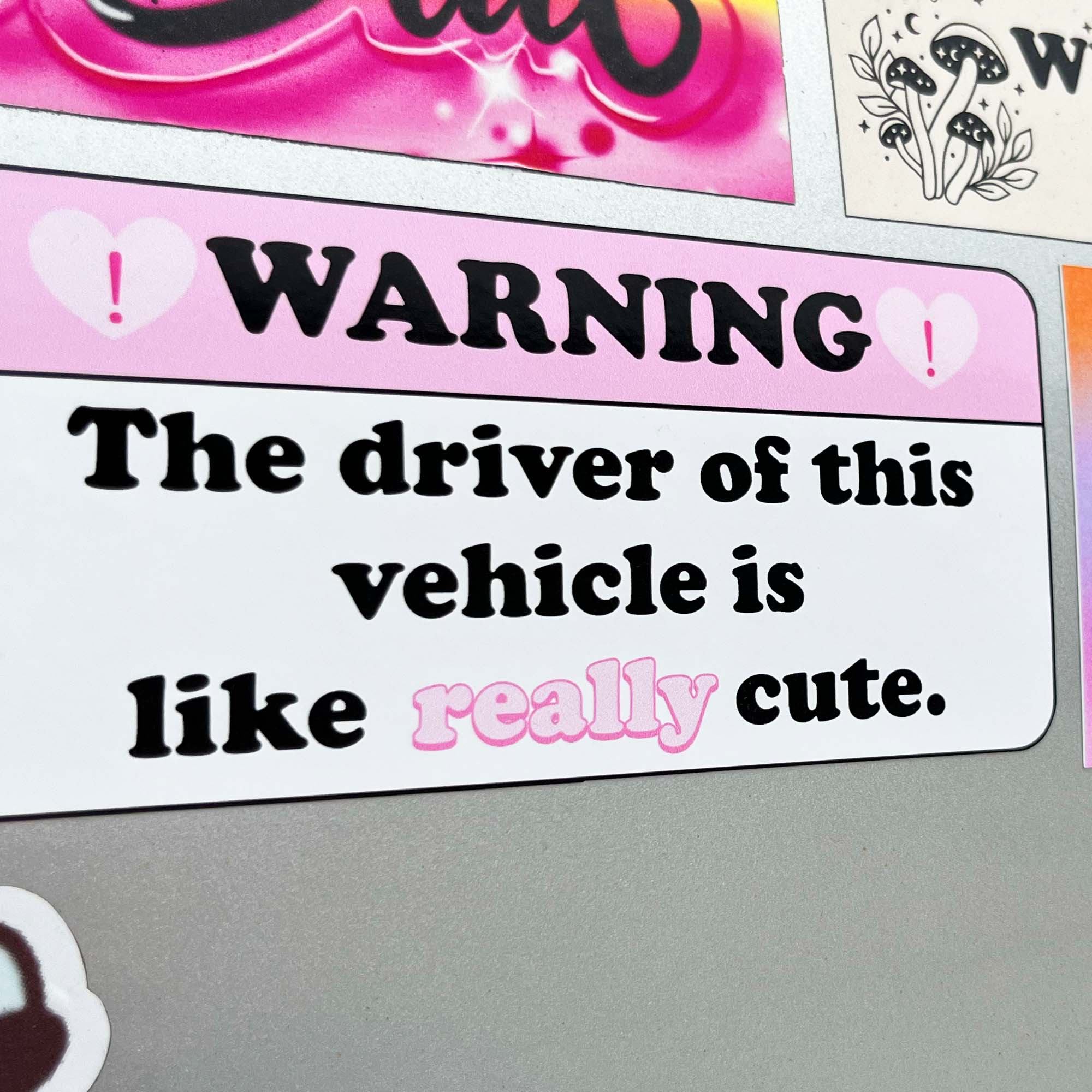 A bumper sticker with a pink background and black text, featuring the phrase 'WARNING The driver of this vehicle is like really cute.'.