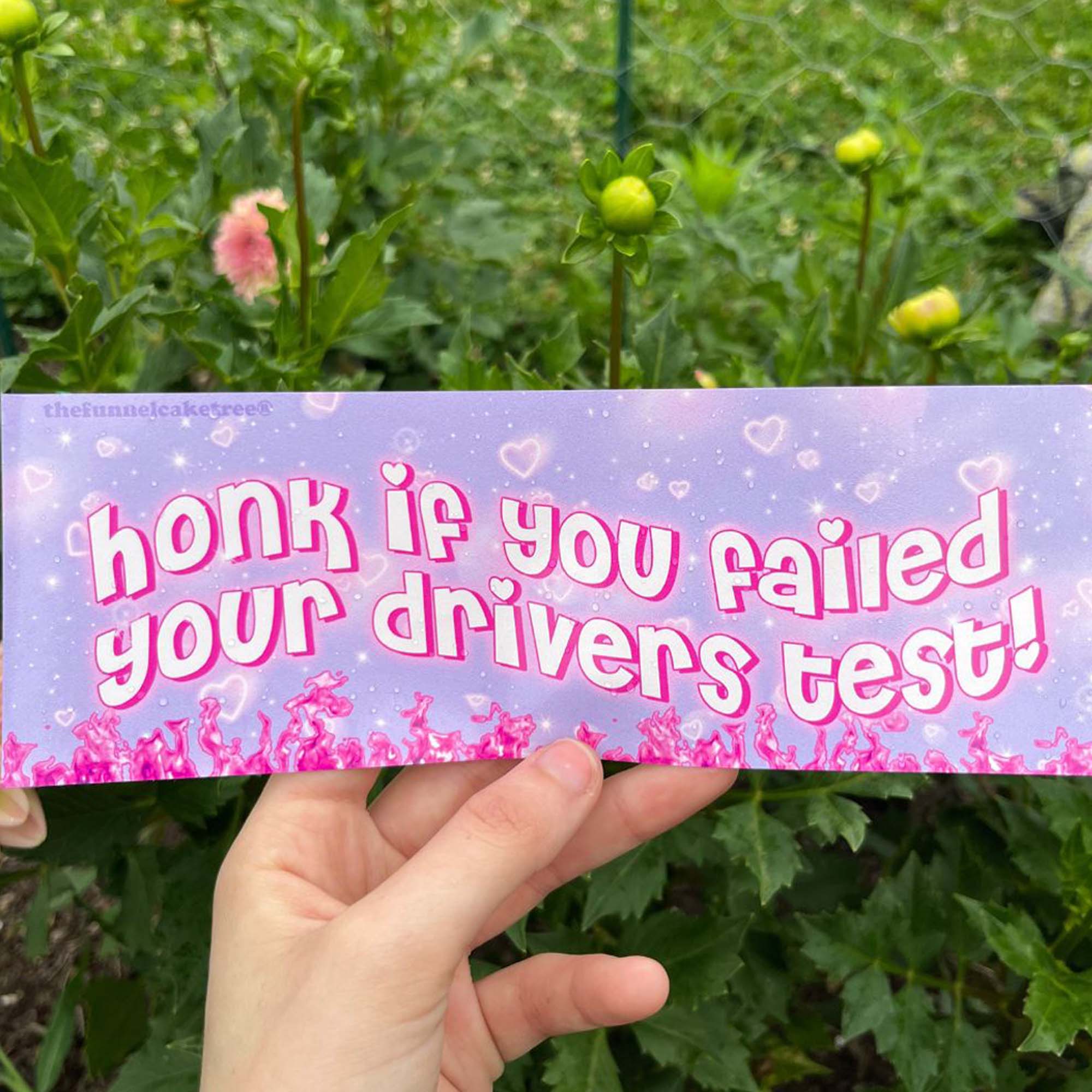 A hand holding a bumper sticker with the text 'honk if you failed your drivers test' on it, surrounded by greenery in the background.