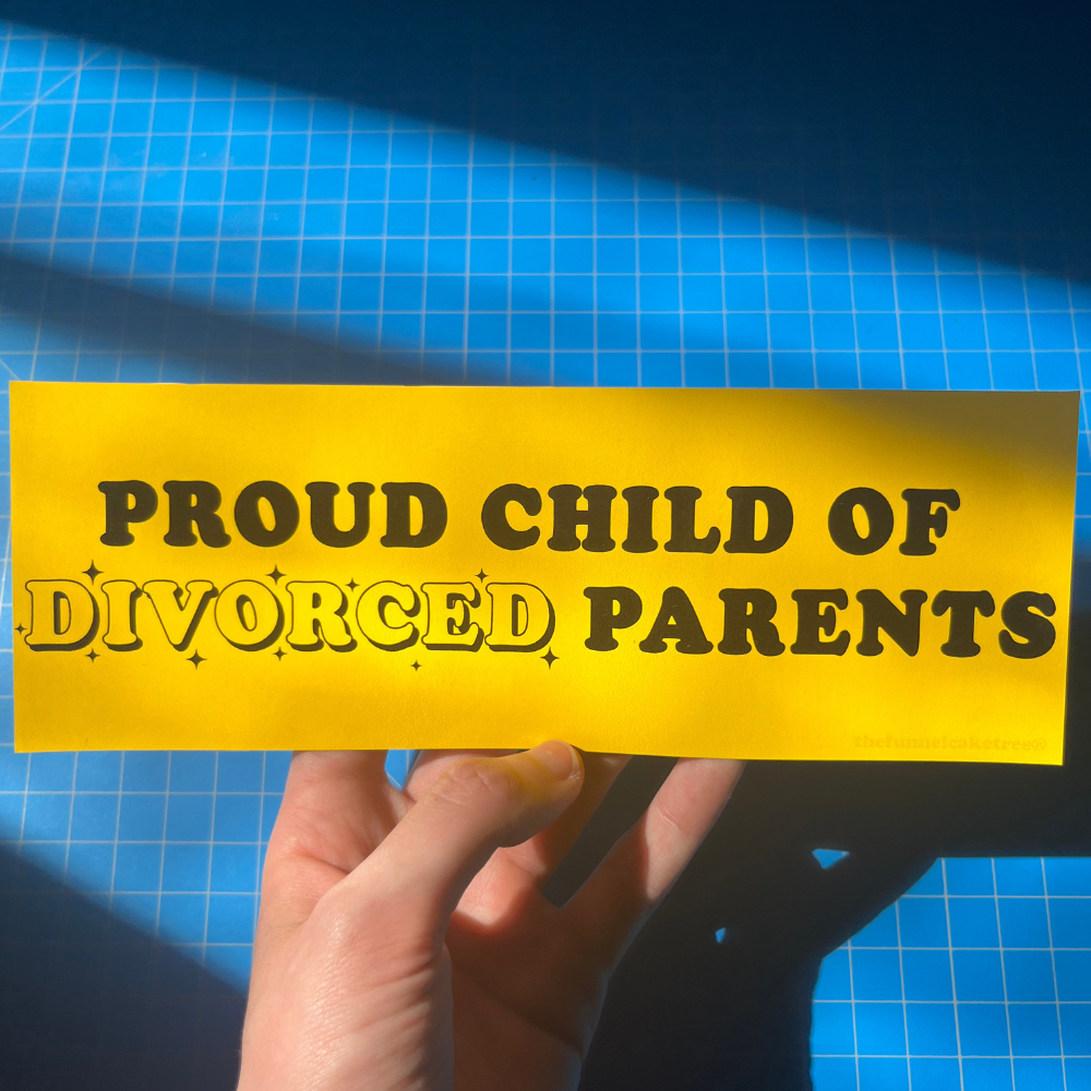A hand holding a yellow bumper sticker with the text 'PROUD CHILD OF DIVORCED PARENTS' in black capital letters.